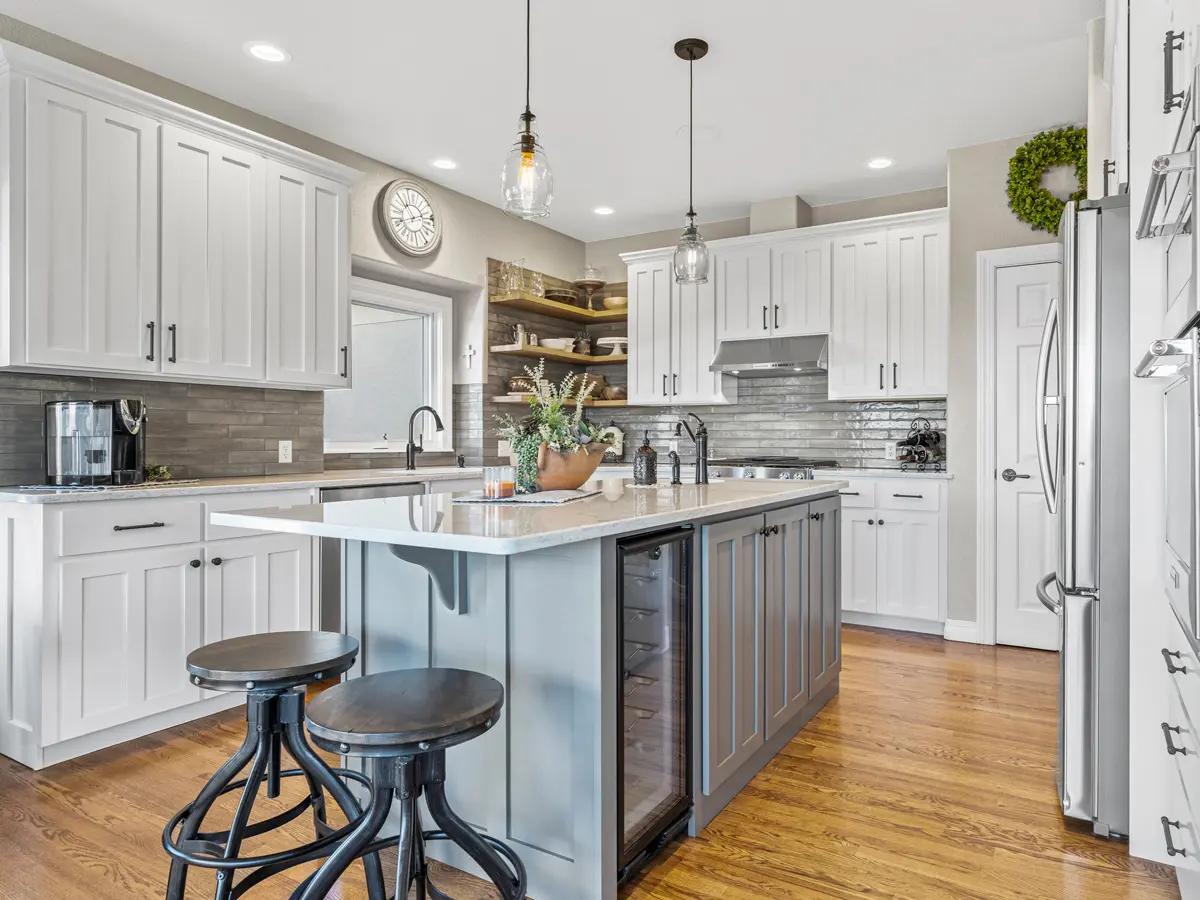 Spacious kitchen with white cabinetry, gray island, and wood floor. Features pendant lights, barstools, and modern appliances. Bright, welcoming tone.