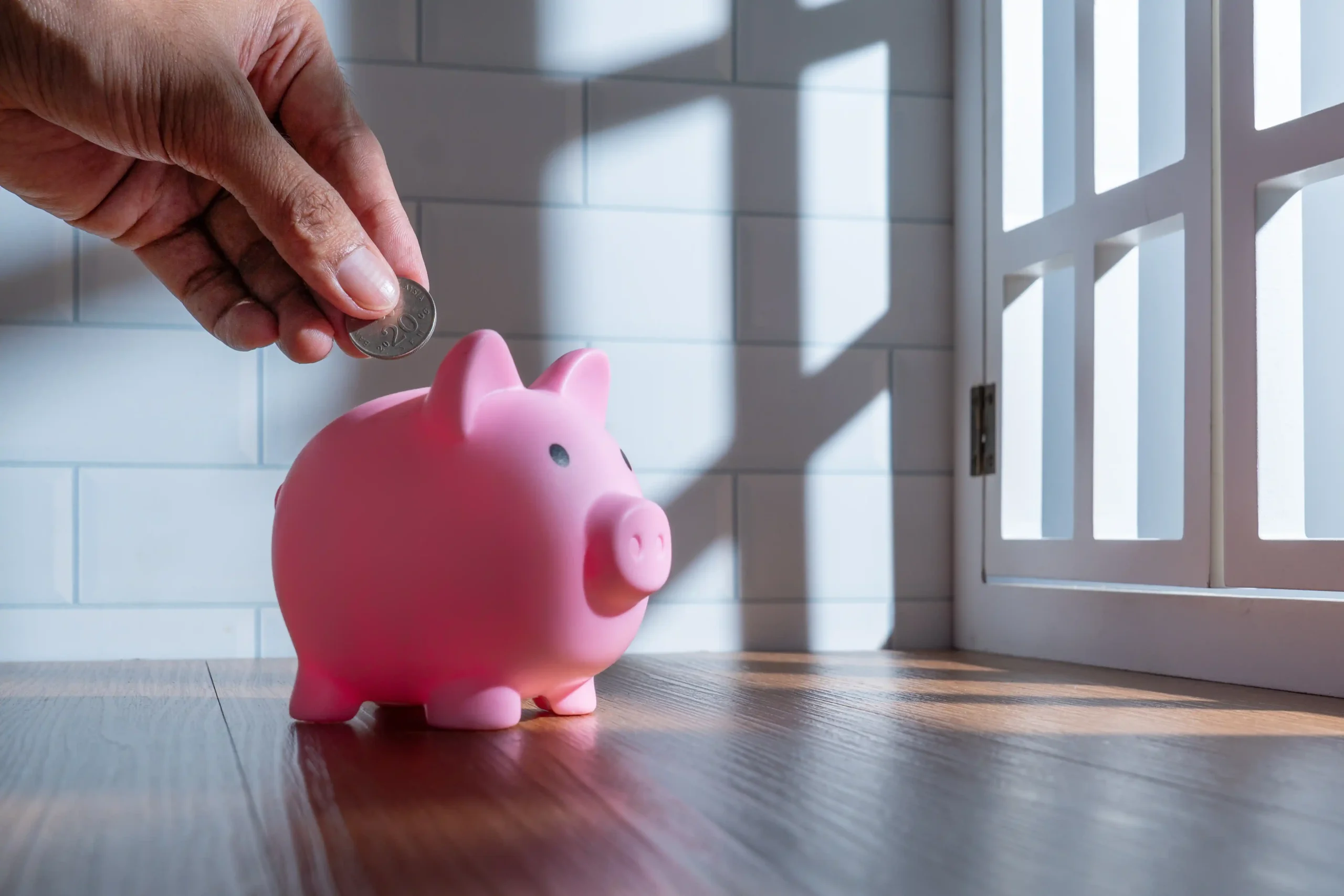 A hand places a coin into a pink piggy bank on a sunlit wooden floor. Light streams through a window, creating a warm, hopeful atmosphere.