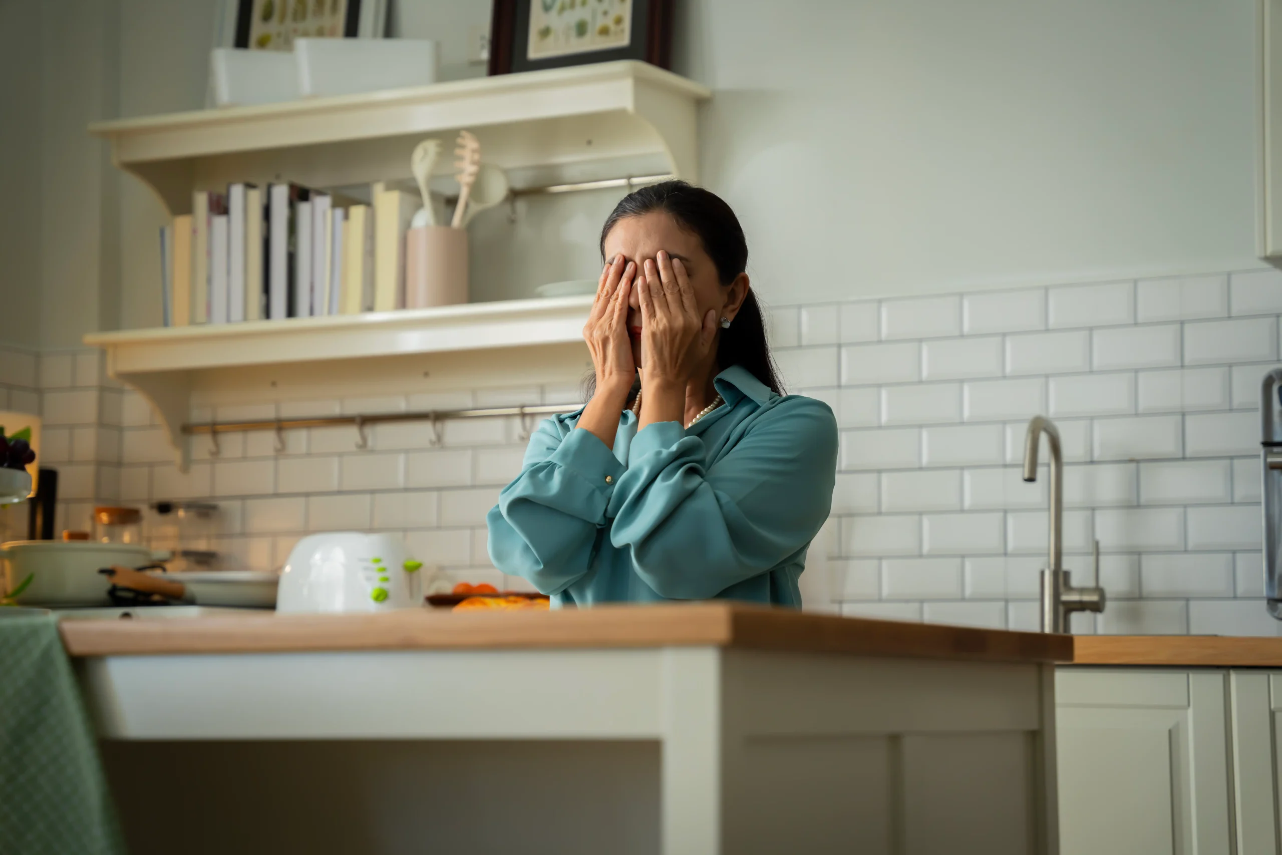 A woman in a teal blouse stands in a kitchen, hands covering her face. The surroundings are bright, with white tile backsplash and open shelves. She appears stressed.