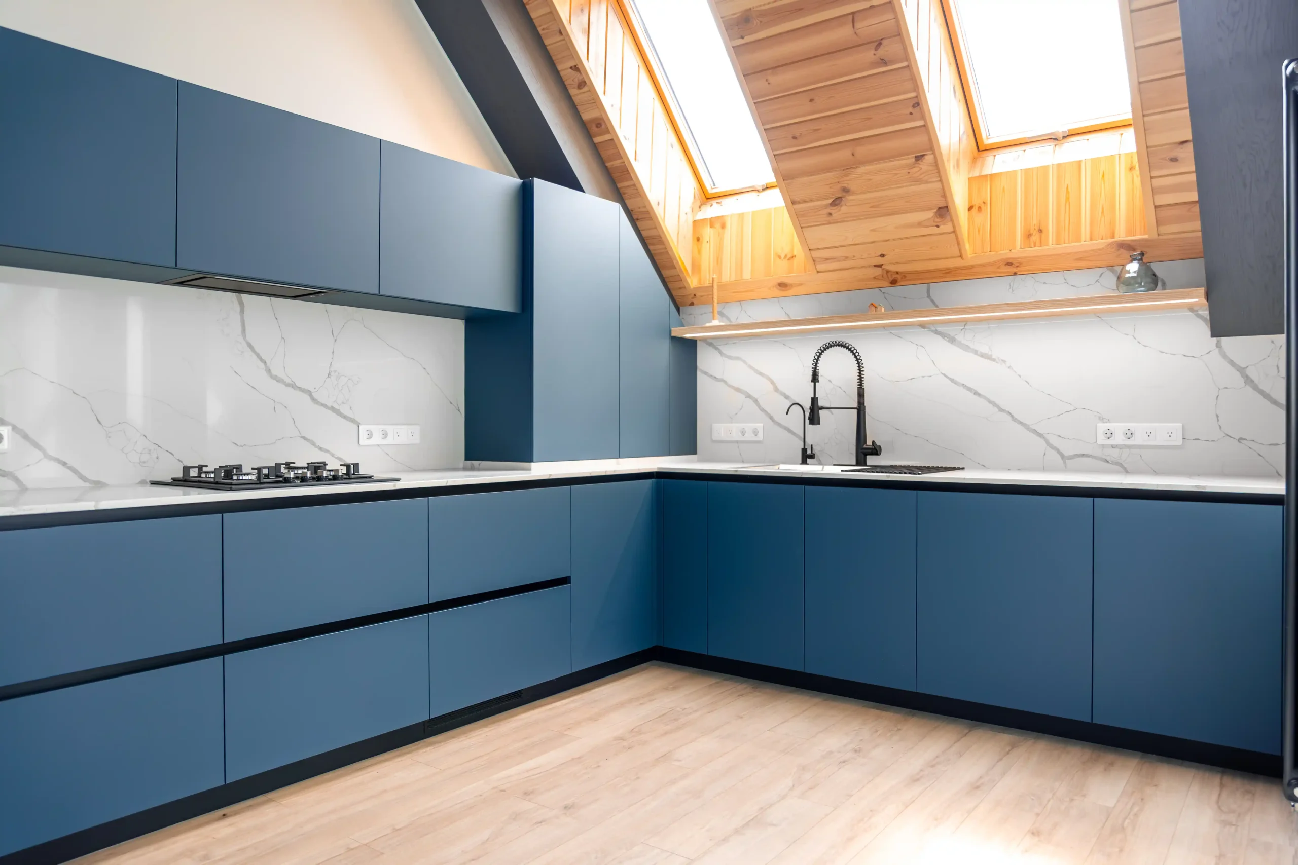 Modern kitchen with sleek blue cabinets under a bright, sloped wooden ceiling. White marble backsplash, black faucet, light wood flooring. Cozy and airy.