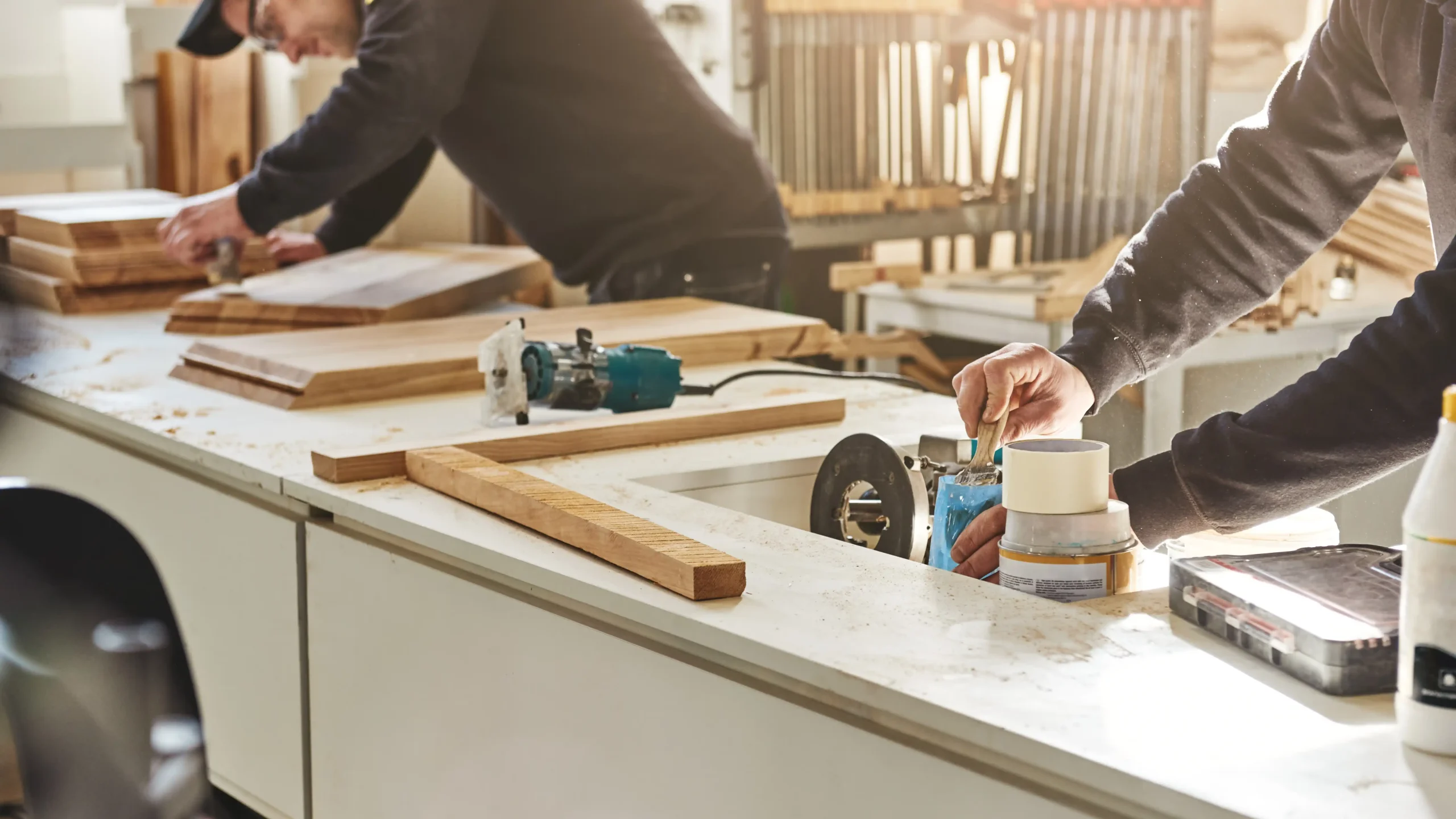 Two people work in a sunlit workshop, crafting wooden pieces on a table. Tools and wood shavings are visible, creating a focused, industrious atmosphere.