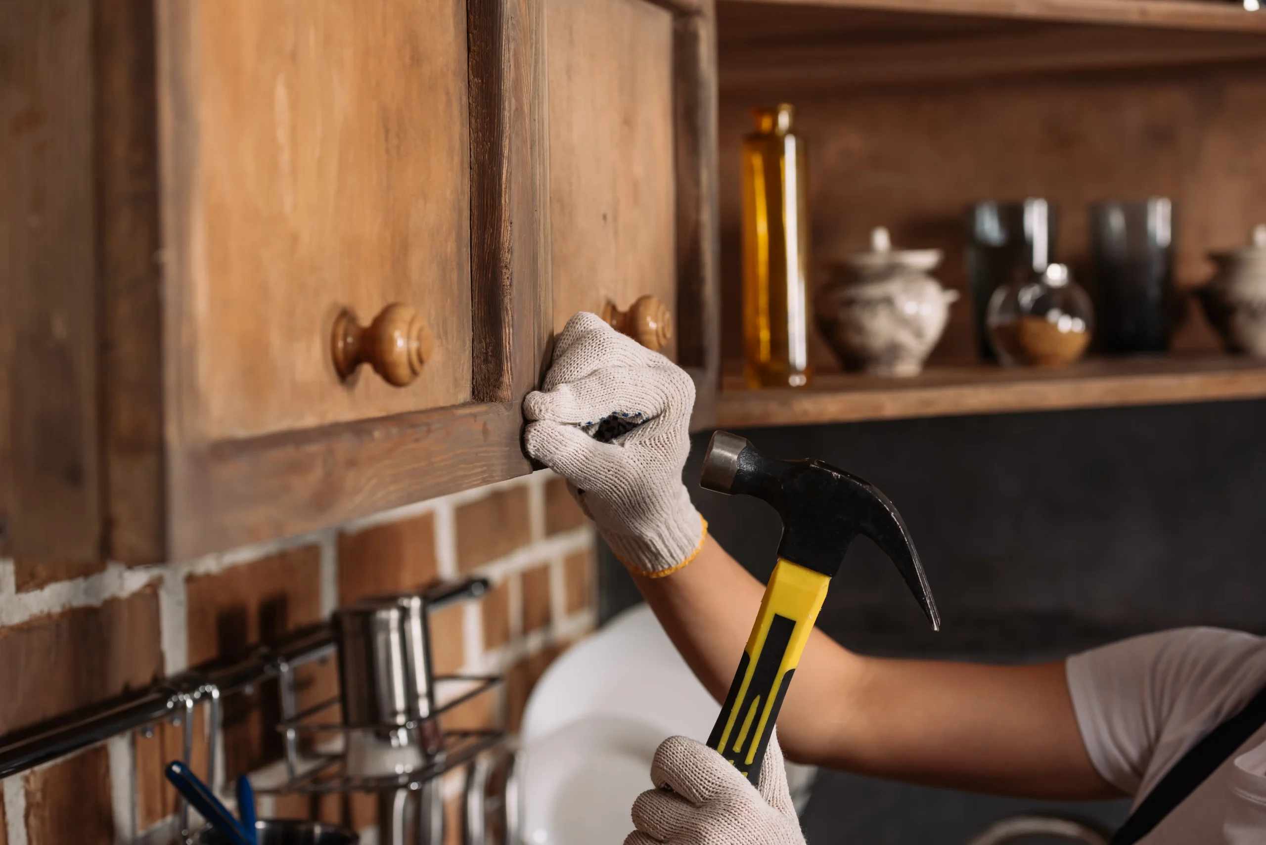 A person wearing gloves uses a hammer to fix a wooden kitchen cabinet, suggesting a home repair or DIY project. Shelves with jars are visible.