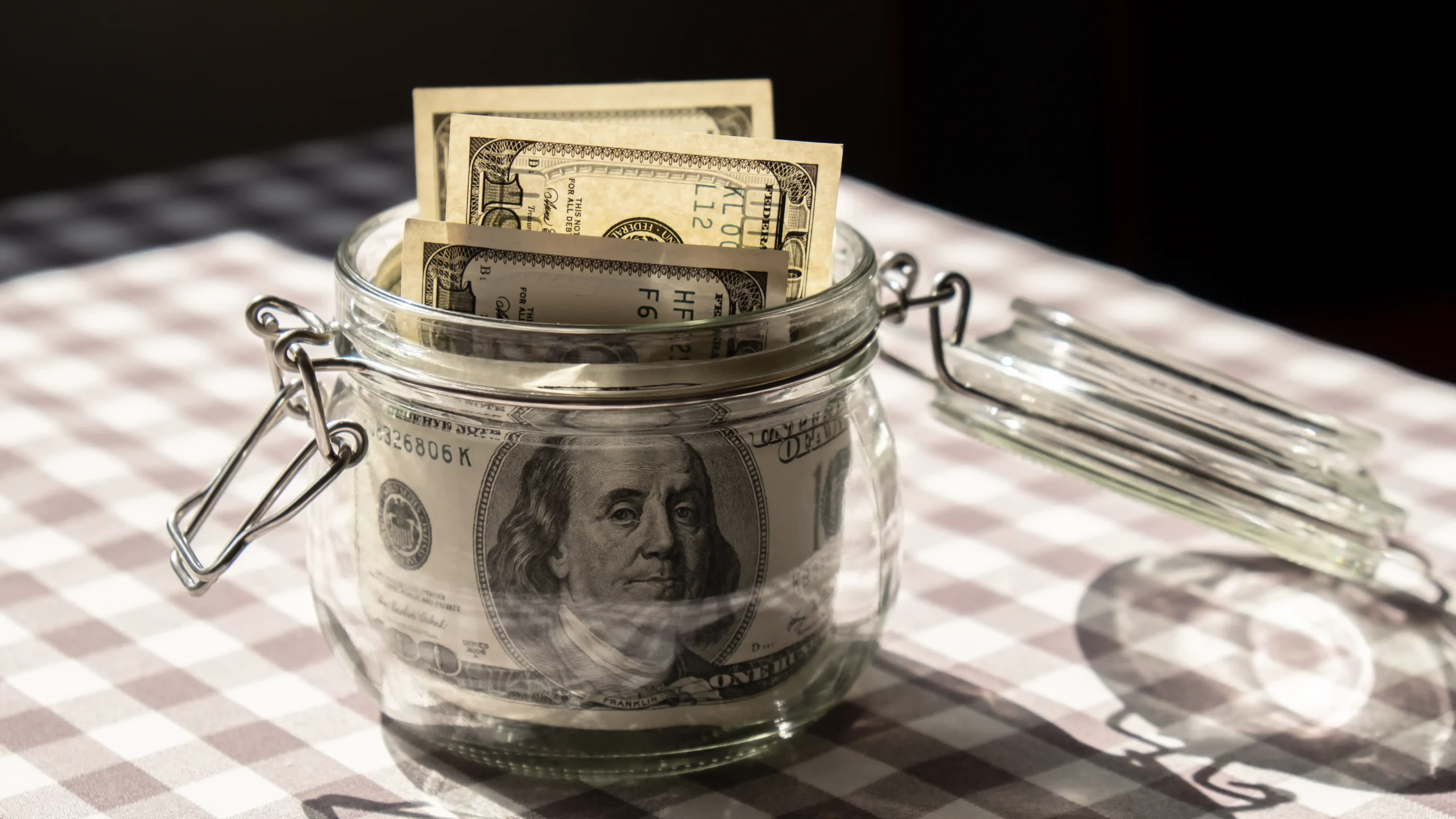 A glass jar sits on a plaid tablecloth, filled with various denominations of U.S. dollar bills, suggesting savings or tip collection. Warm lighting casts soft shadows.