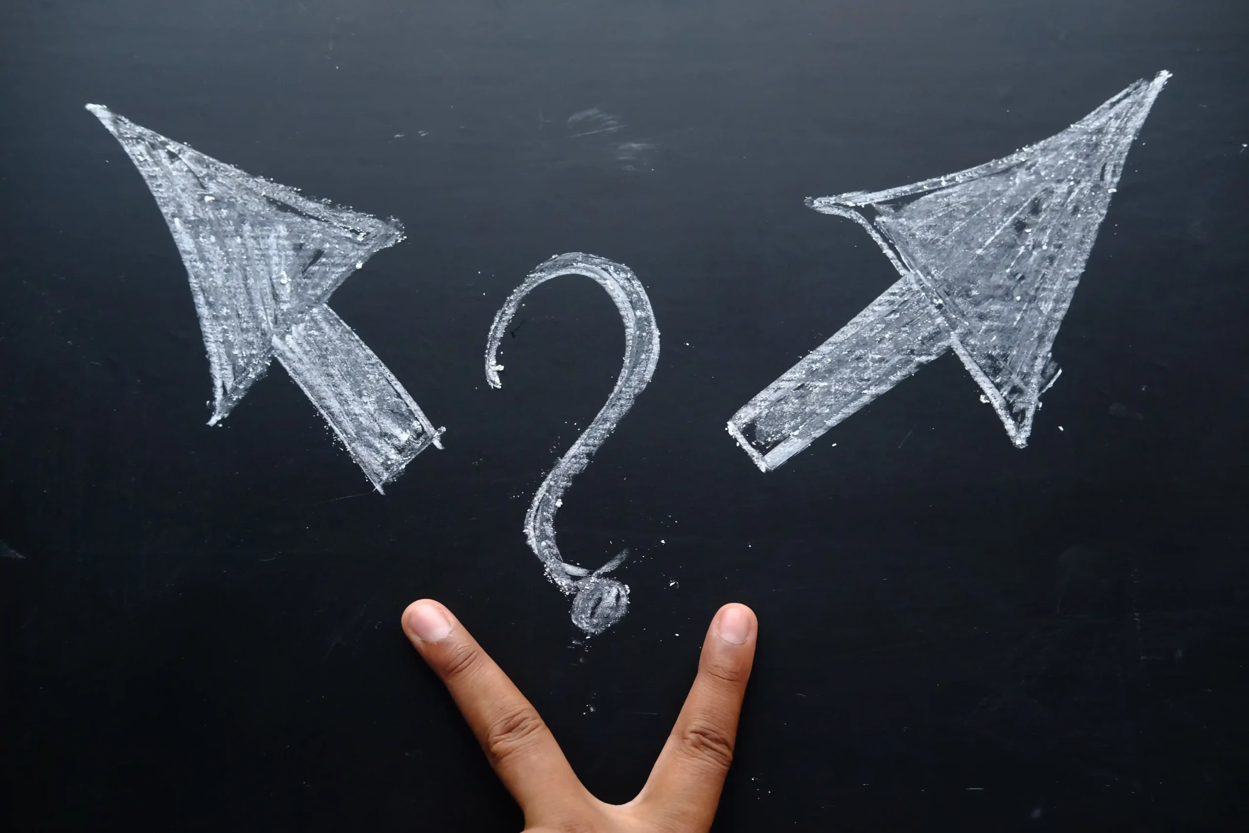 A hand forms a peace sign in front of a chalkboard. Above, a chalk-drawn question mark is flanked by two arrows pointing left and right, symbolizing choice.