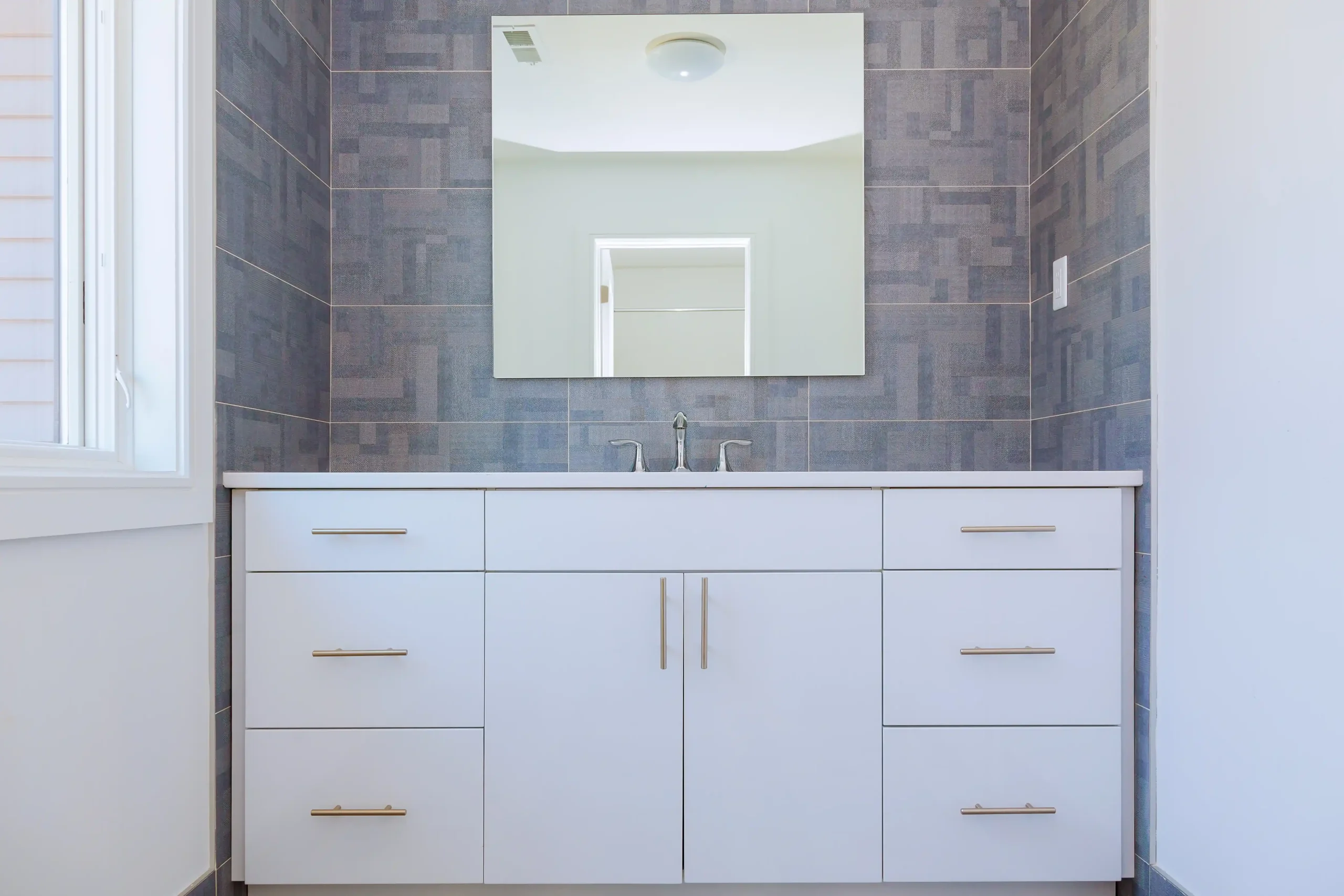Modern bathroom with a white vanity, sleek silver handles, and a large mirror reflecting a doorway. Gray textured tiles add a sophisticated touch.