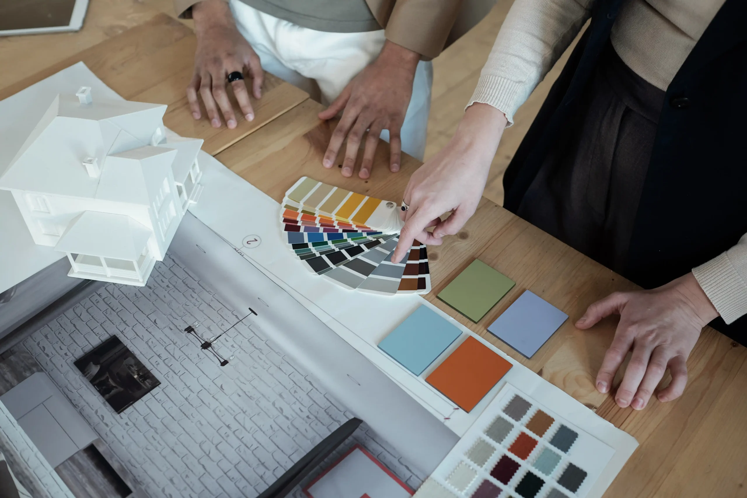 Two people are discussing house design plans, with one pointing at a palette of color swatches. A house model and architectural drawings are visible, conveying creativity and collaboration.