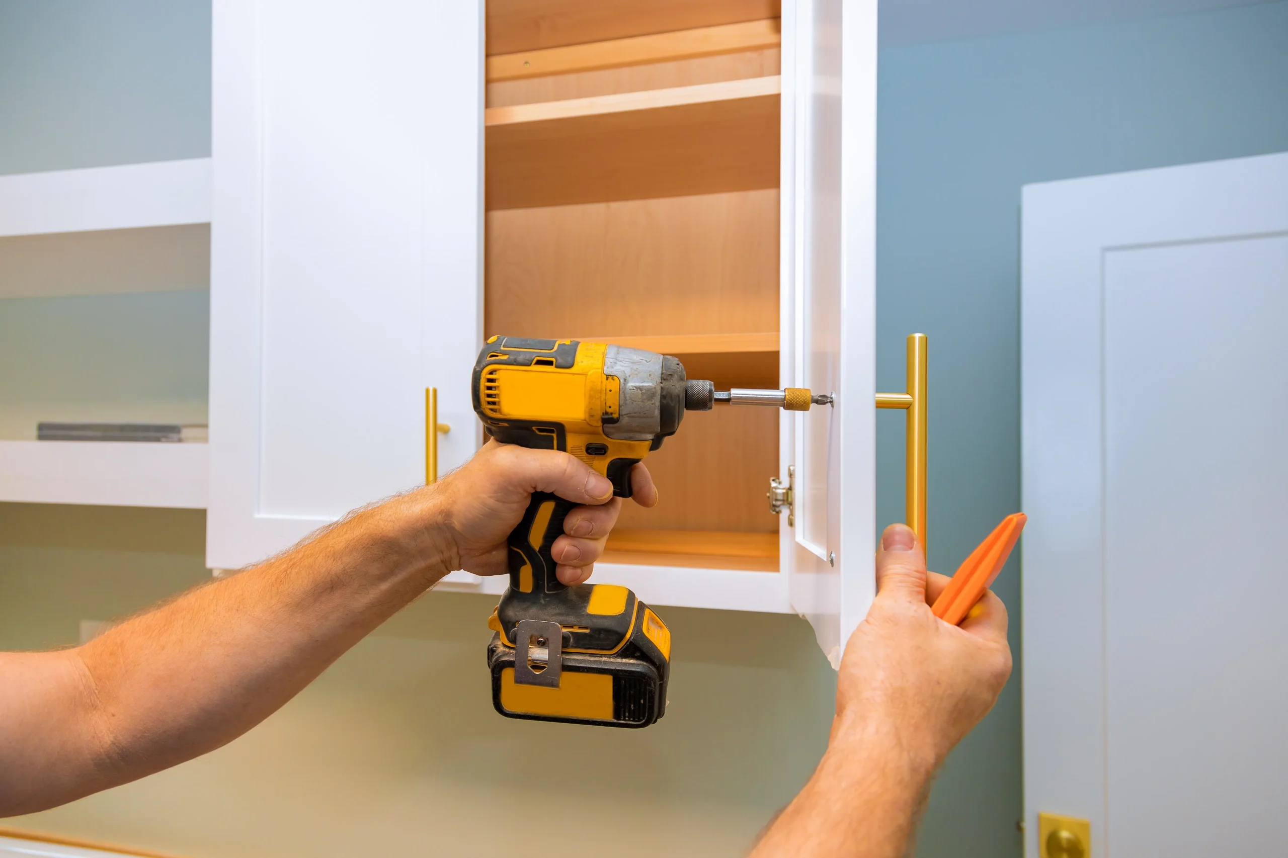 A person uses a yellow drill to install a handle on a white cabinet door, with focus and precision. The cabinet is open, revealing wooden shelves inside.