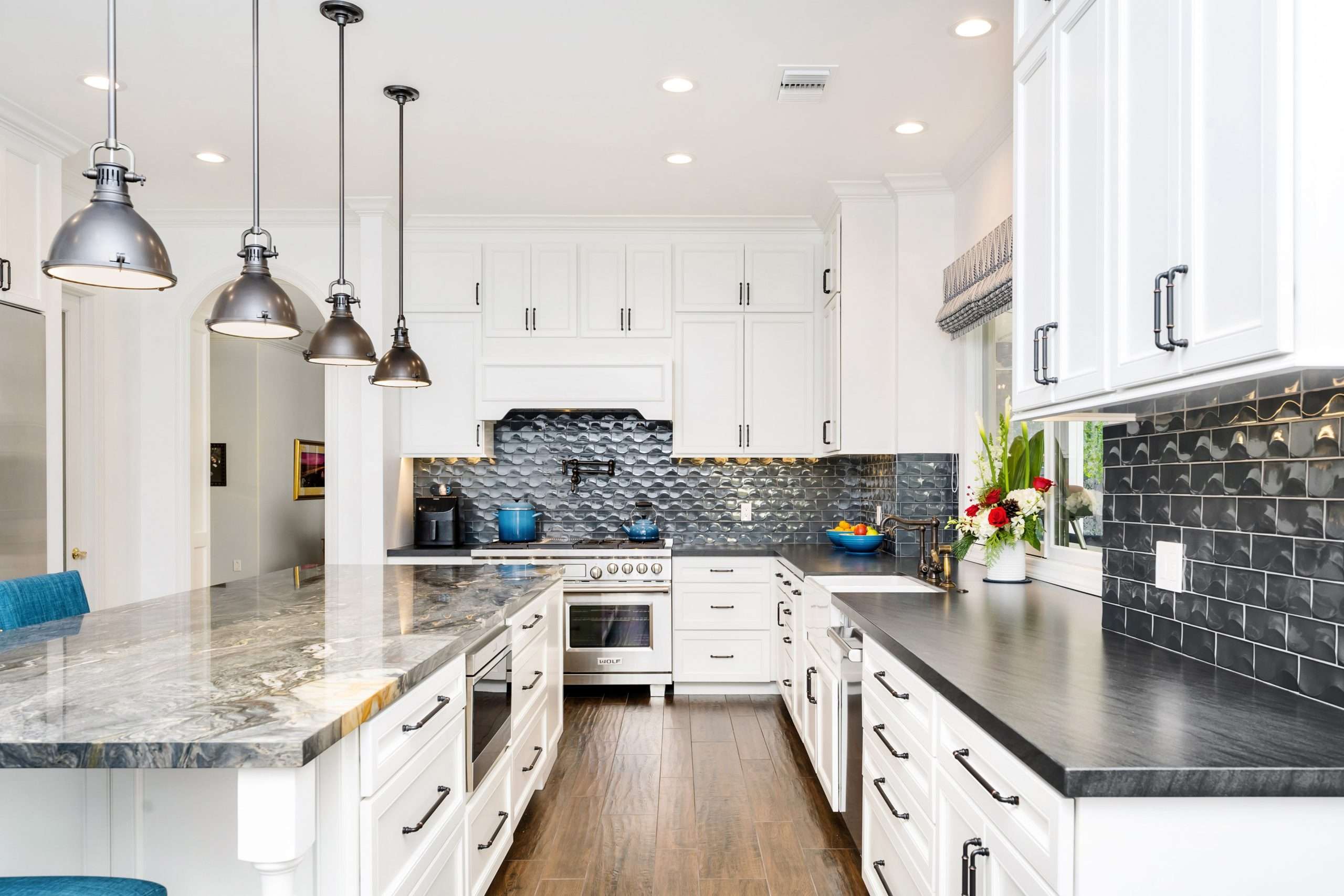 Bright kitchen with white cabinets, dark countertops, and a glossy black tiled backsplash. Pendant lights hang over a large island. Elegant and modern.