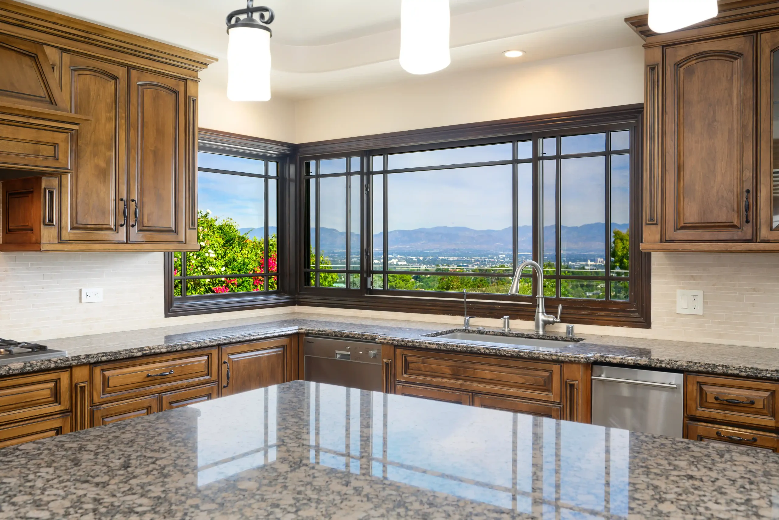 Spacious kitchen with wooden cabinets, granite countertops, and a large window with a scenic mountain view. Bright pendant lights illuminate the area.