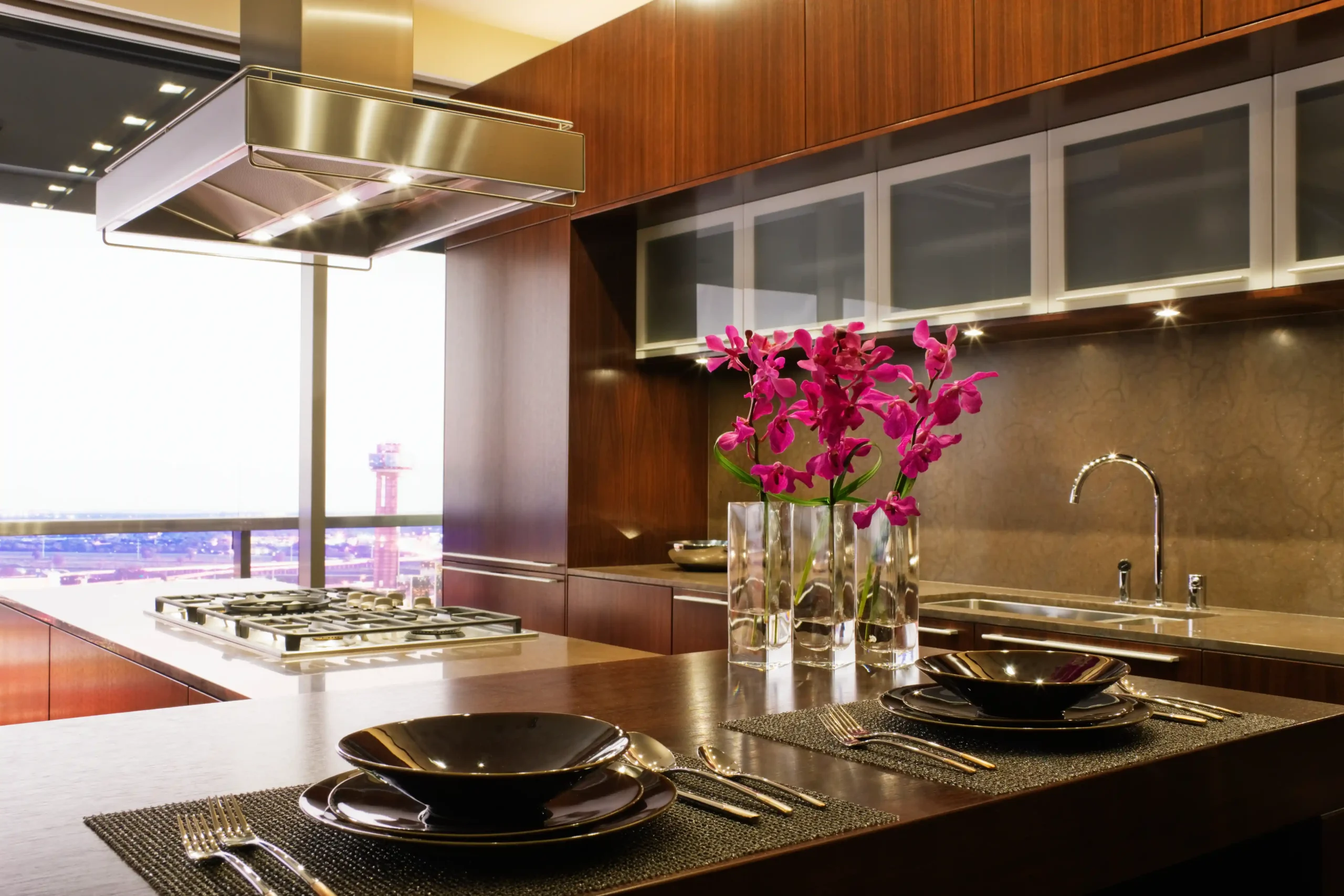 Modern kitchen with sleek wood cabinetry, a gas stovetop under a steel hood, vibrant pink flowers on the counter, and elegant table settings.