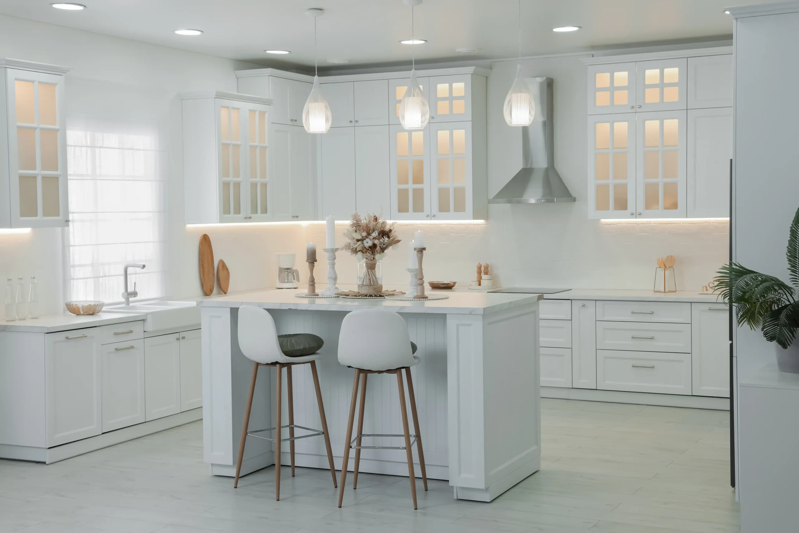 Sleek white kitchen with a central island, two stools, elegant pendant lights, glass cabinets, and subtle plant decor, creating a serene atmosphere.