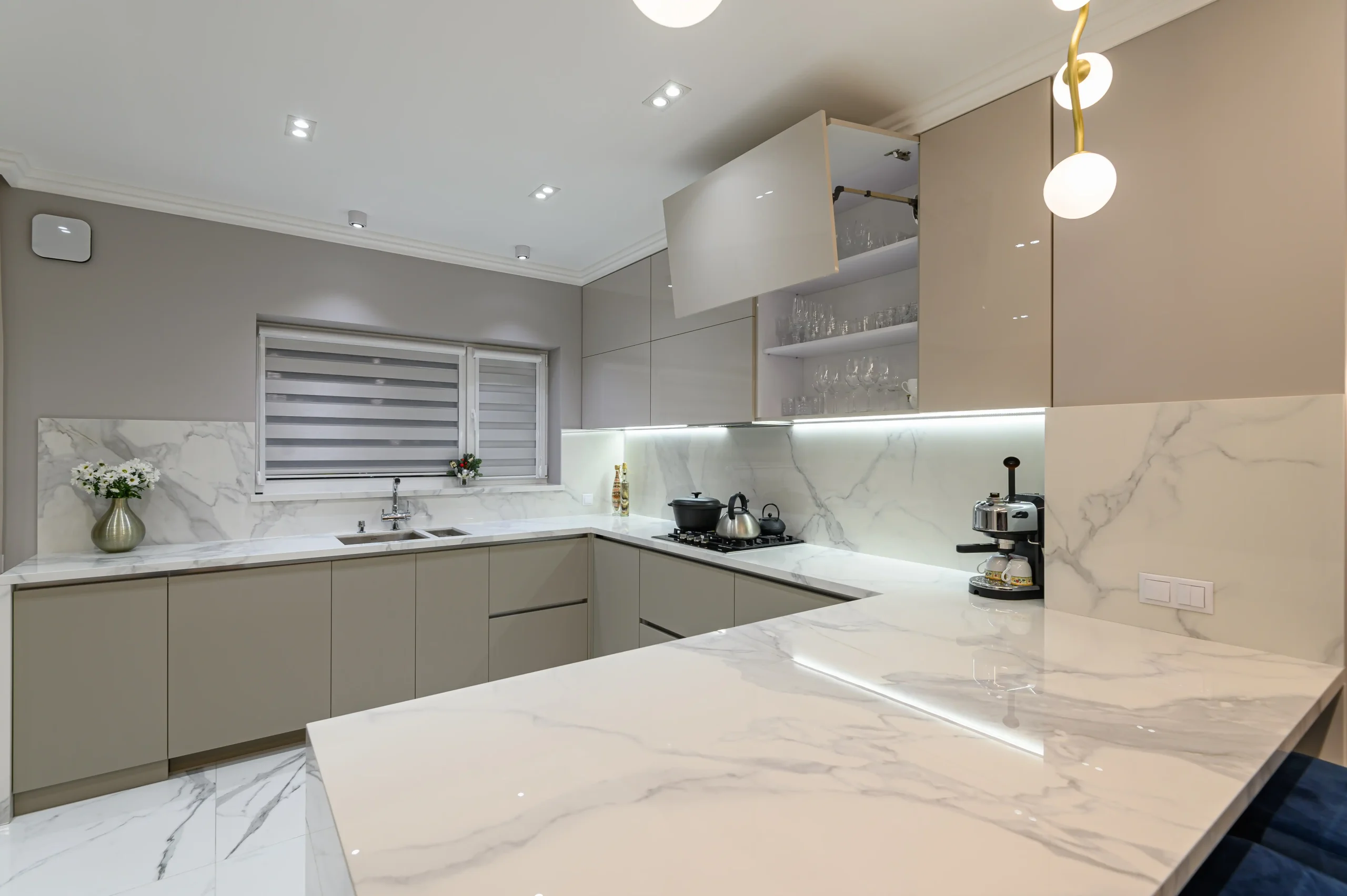 Modern kitchen with sleek beige cabinets, marble countertops, and backsplash. Overhead lights brighten the space. A vase, appliances, and open shelf add detail.