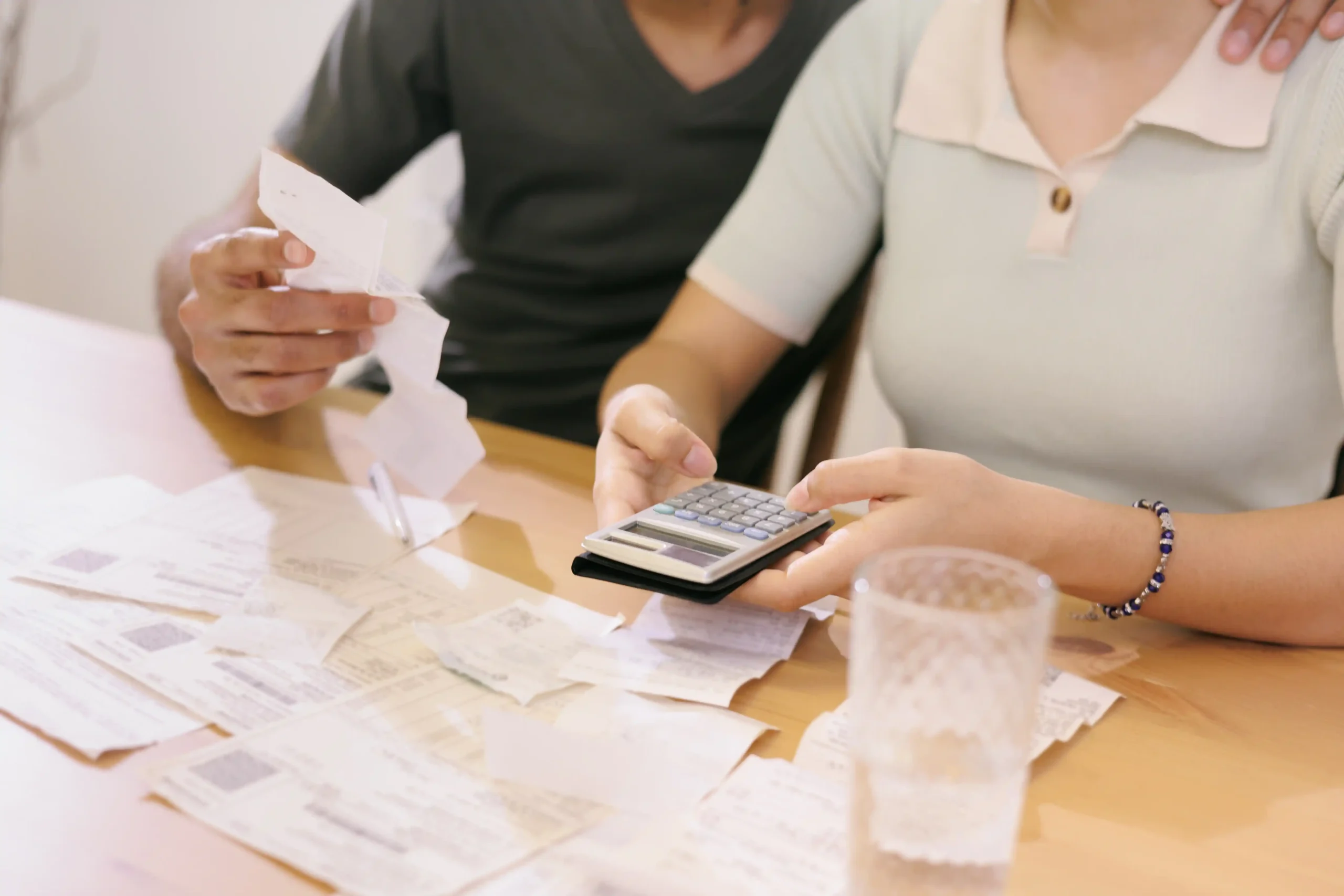 A couple sits at a wooden table covered with scattered bills and receipts. They use a calculator, conveying focus and collaboration in handling finances.