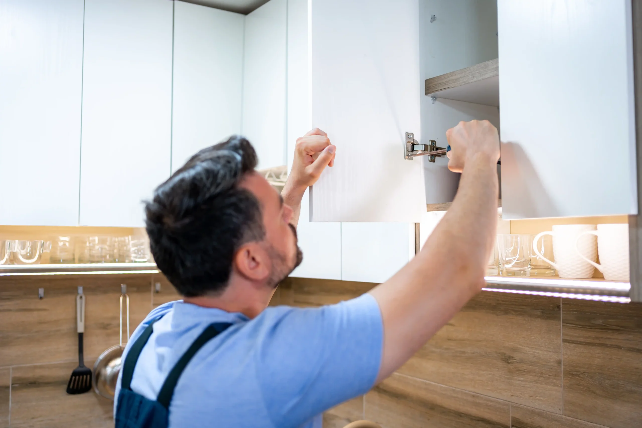 A man in a blue shirt and overalls repairs a kitchen cabinet hinge. The bright kitchen has white cabinets and wooden walls, creating a calm atmosphere.