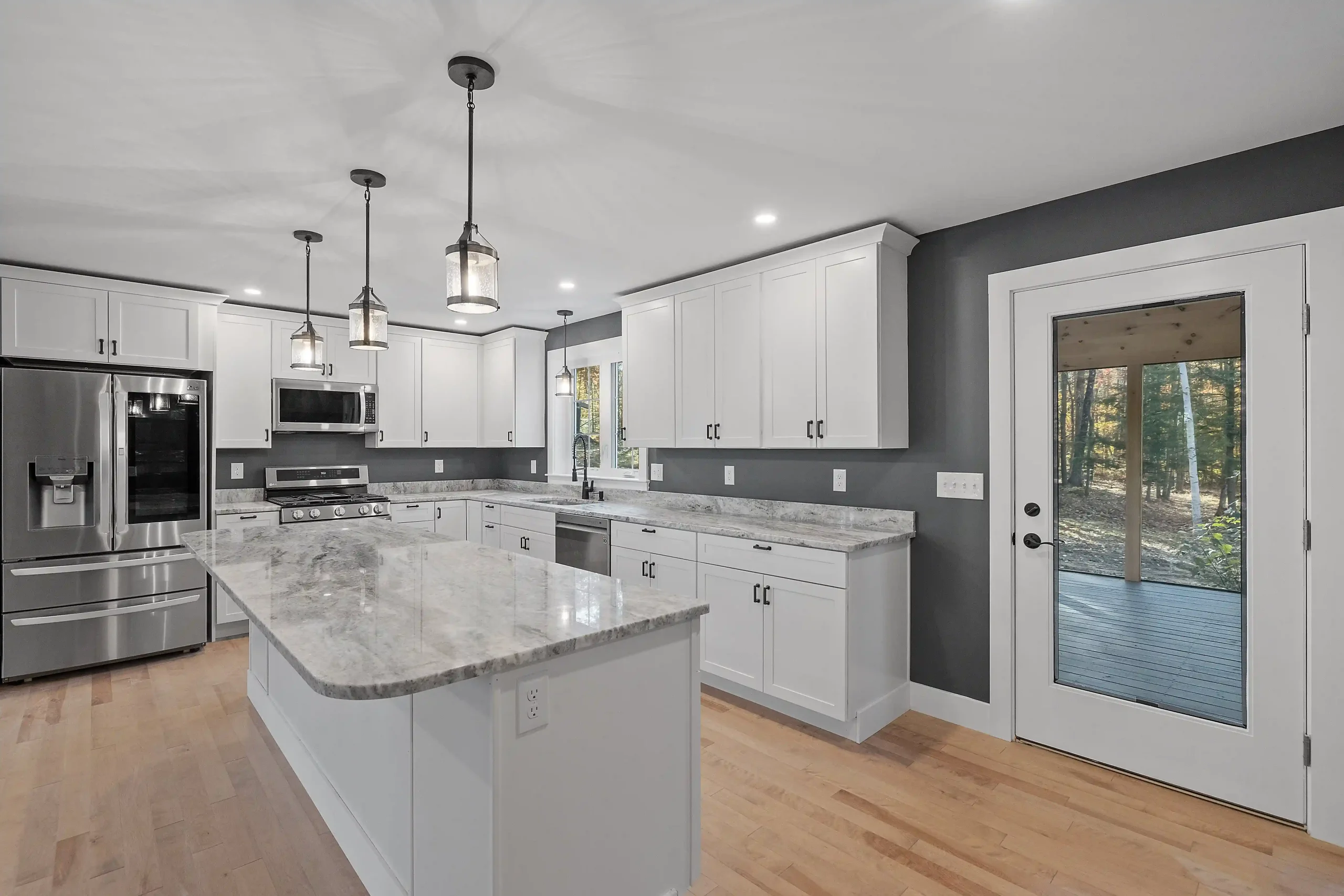 Modern kitchen with white cabinetry and marble countertops. Stainless steel appliances are visible, with pendant lights above. A door opens to a forest view.