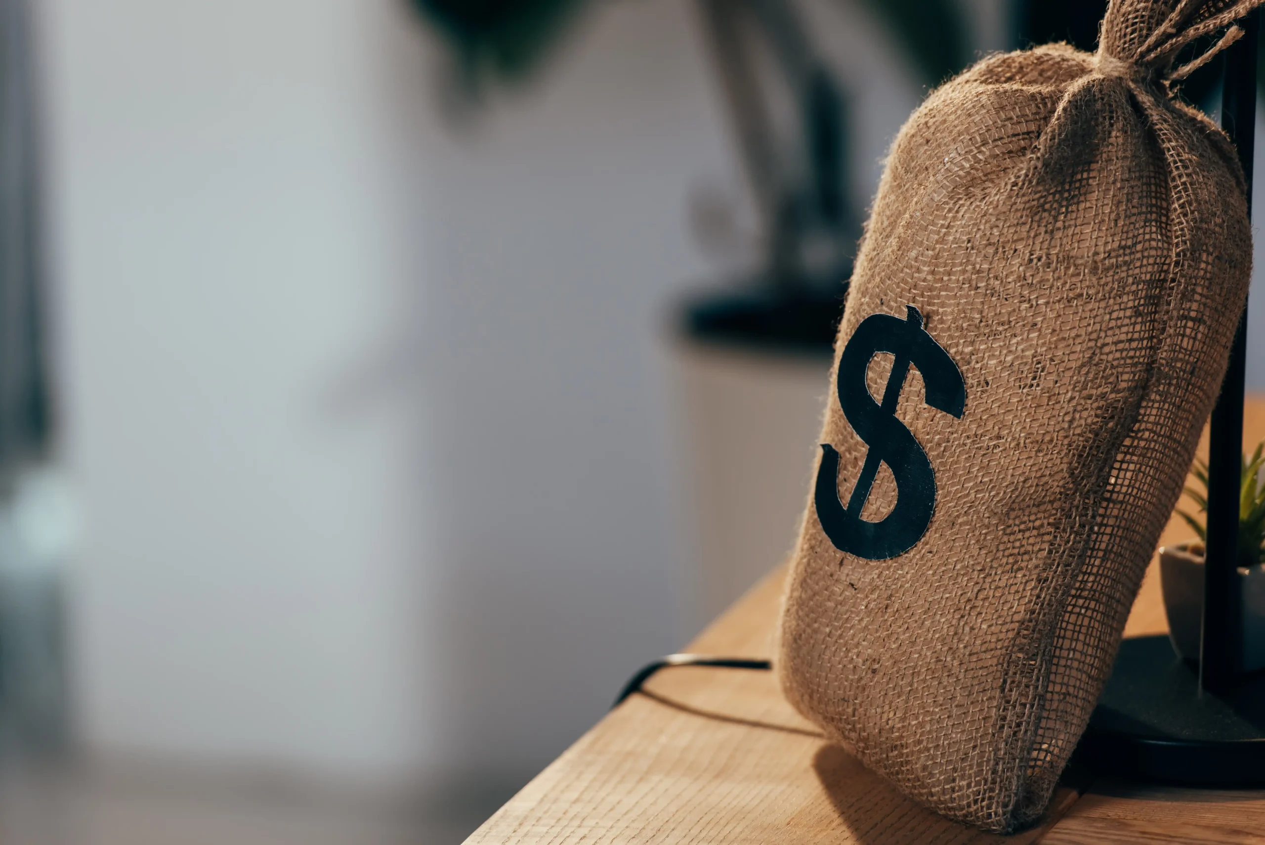 A burlap sack with a black dollar sign is placed on a wooden desk. The background is blurred, creating a calm, focused atmosphere.