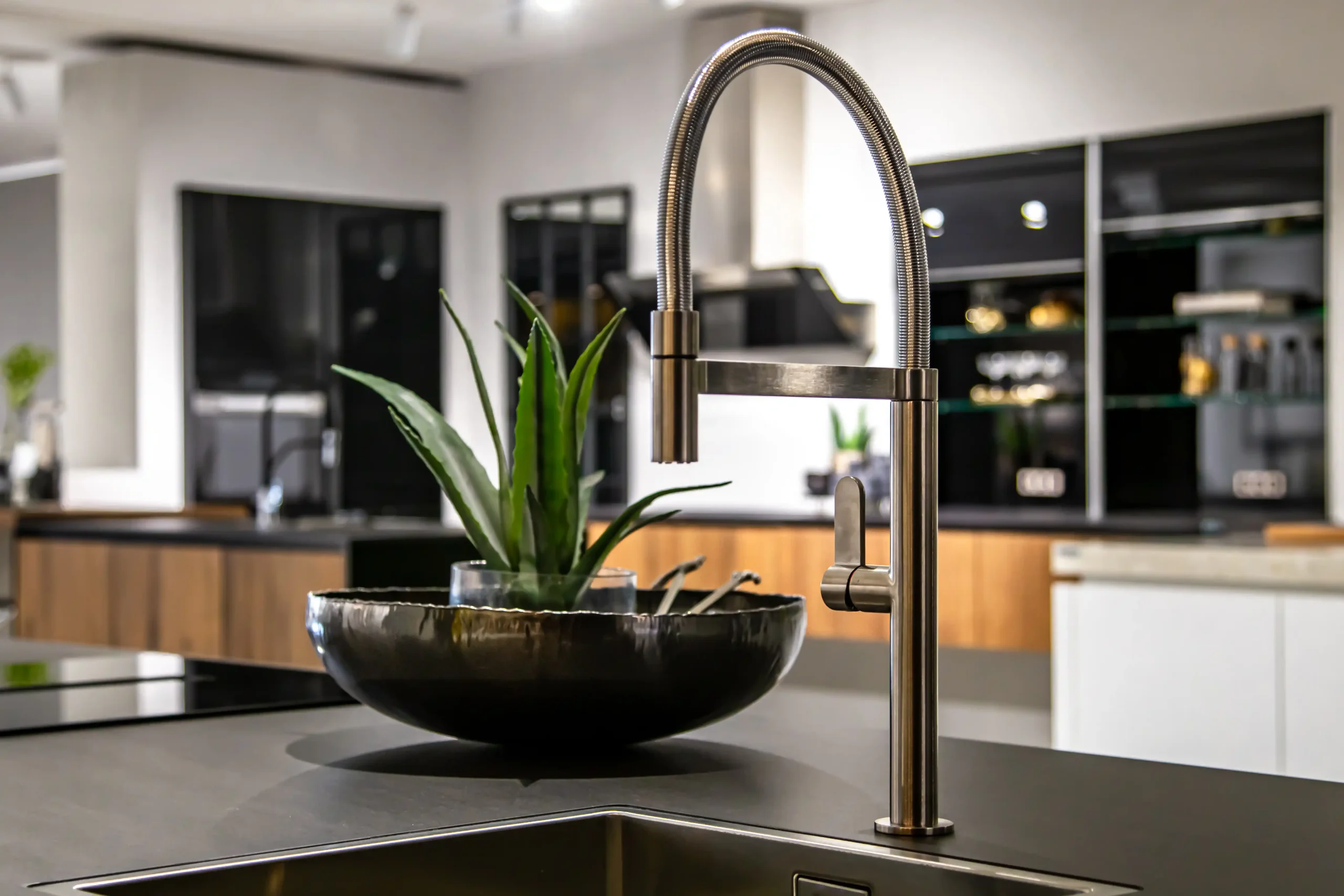 Modern kitchen with a sleek chrome faucet and a green plant in a black bowl on a dark countertop. Background features wooden cabinets and open shelving.