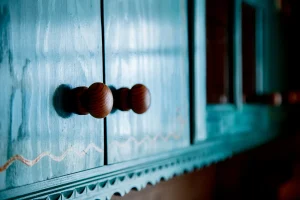 Close-up of an ornate turquoise cabinet with wooden knobs. The cabinet features decorative scalloped edging and a subtle wavy pattern, evoking a rustic charm.