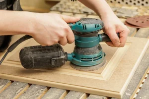 Hands using an electric sander on a wooden board, with fine wood dust visible. The scene conveys focus and craftsmanship in woodworking.