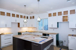 Modern kitchen under construction with white cabinetry, central island, and pendant lights. Counters covered for protection, tools visible, bright atmosphere.