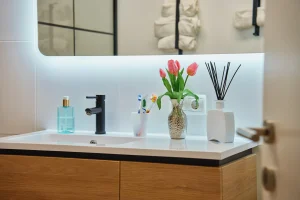 Modern bathroom vanity with wooden drawers, a sleek black faucet, toothbrush holder, pink tulips in a vase, and a diffuser, exuding a fresh, elegant vibe.