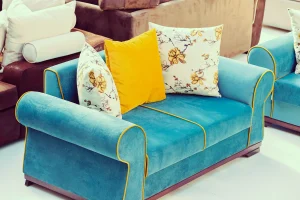 A vibrant blue sofa with yellow piping, adorned with two floral pillows and a bright yellow pillow, sits in a cozy, colorful showroom setting.