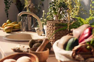 Sunny kitchen counter with fruits, vegetables, and plants. A wicker basket, bananas, kiwis, and potted herbs create a fresh, inviting atmosphere.