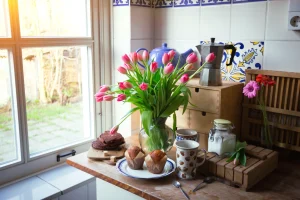 A kitchen counter with a vase of pink tulips, muffins on a plate, a chocolate cake, coffee maker, cups, and a jar of sugar. Sunlight streams through a window.