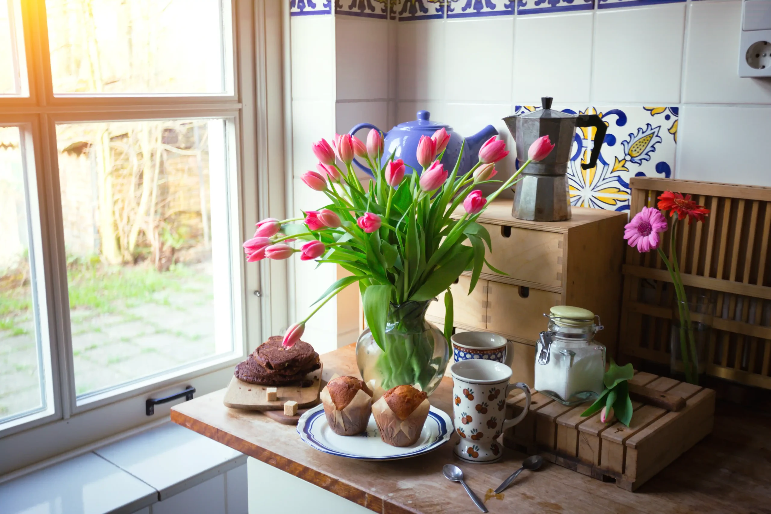 A kitchen counter with a vase of pink tulips, muffins on a plate, a chocolate cake, coffee maker, cups, and a jar of sugar. Sunlight streams through a window.