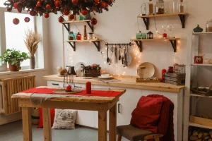 Cozy kitchen decorated for Christmas with hanging garland, red ornaments, and candles. Wooden shelves hold jars and festive items. Warm, inviting ambiance.